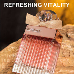 A rectangular glass perfume bottle with a rose gold cap and a small beige bow sits on a rock.