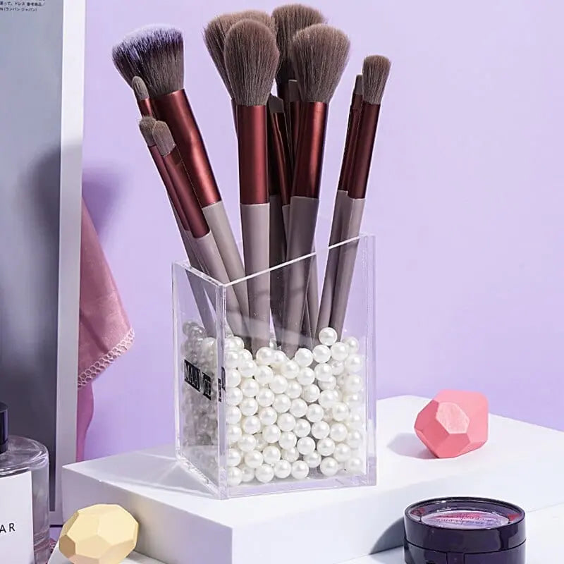 A clear acrylic container filled with white pearls holds several brushes from the 13 PCS Makeup Brushes Set Eye Shadow Foundation Women Cosmetic Brush—displayed near pastel decorations and a small round makeup product on a white surface.