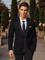 A man wears a black three-piece suit with a white shirt and black tie in an outdoor setting.