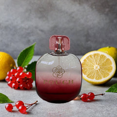 A bottle of Pouch Blush Rose perfume sits on a grey surface surrounded by lemons and red berries.