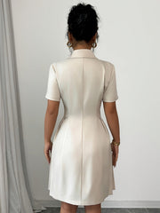 A woman wears a light beige, short-sleeved, A-line dress with a lapel collar from the back.