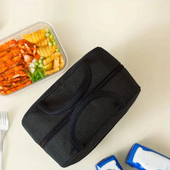 A black, rectangular insulated lunch bag with top handles sits on a table next to a meal container.
