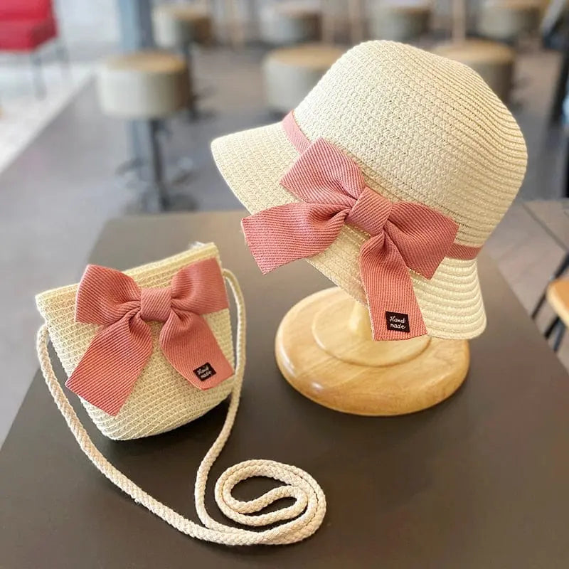 A 2-8 Years Kids Sun Hat Girls Summer New Straw Bag Cap Set, featuring a beige straw hat with a pink bow on a wooden stand and a matching crossbody bag, is displayed on a table with blurred stools and tables in the background.