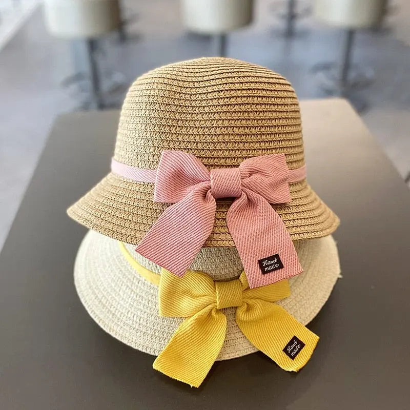 Two kids' summer straw hats from the Sun Hat & Bag Set are stacked on a table. The top hat is light brown with a pink bow, the bottom is beige with a yellow bow, and both have small black tags attached. Suitable for ages 2-8 years.