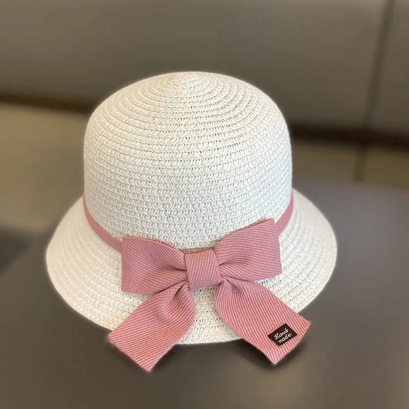 This kids' sun hat is a white straw cap for ages 2-8, featuring a pink ribbon with a large bow and black tag at the back. Ideal for girls, it's perfect for travel or beach days, offering sun protection and style.