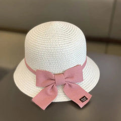 This kids' sun hat is a white straw cap for ages 2-8, featuring a pink ribbon with a large bow and black tag at the back. Ideal for girls, it's perfect for travel or beach days, offering sun protection and style.