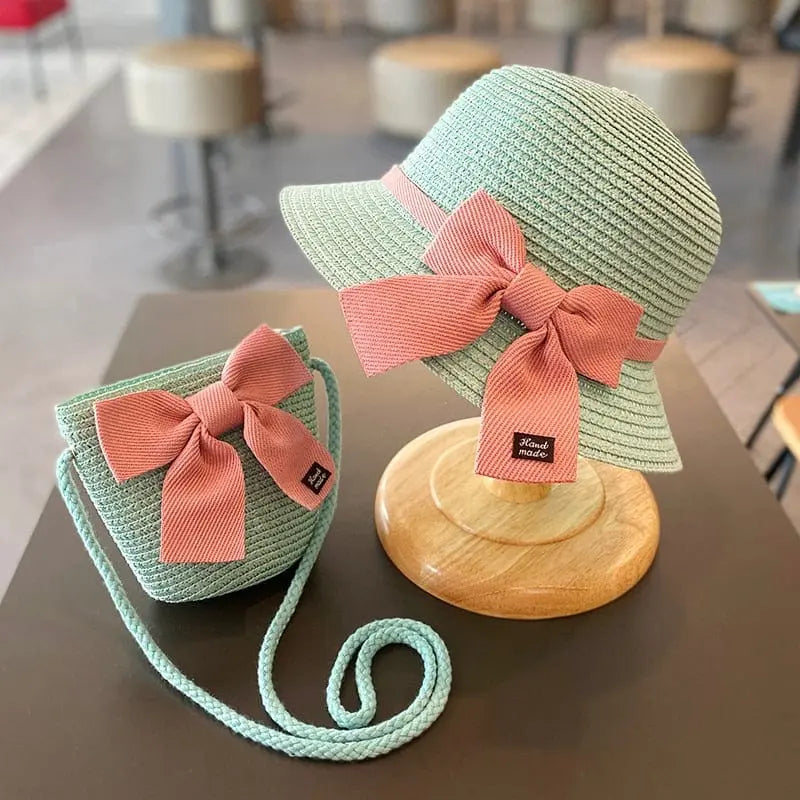 The 2-8 Years Kids Sun Hat Girls Summer New Straw Bag Cap Set features a light green straw beach hat with a wide pink bow and a matching purse with a long strap, both displayed in a cozy cafe setting.