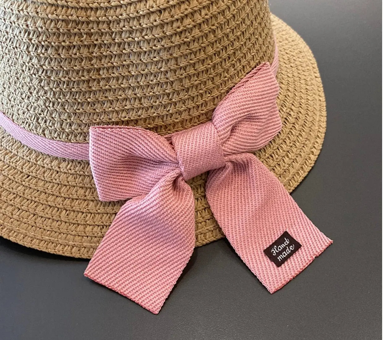 The 2-8 Years Kids Sun Hat is a brown floral straw hat with a pink ribbon bow and "Hand made" black tag, perfect for travel and sun protection. Ideal for girls in summer, this beach fisherman hat is part of a stylish cap set.