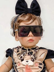 A toddler wearing oversized black square sunglasses with a large black bow headband.