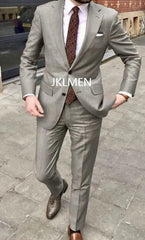 A man wears a light grey two-piece suit with a patterned tie and brown leather shoes.