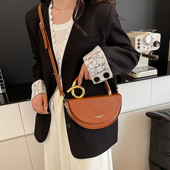 A woman wears a brown saddle-style crossbody bag with a unique gold circular handle.