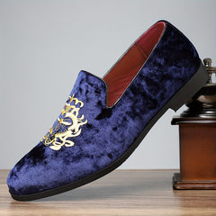 A blue velvet slip-on loafer with gold embroidery resting on a wooden surface.