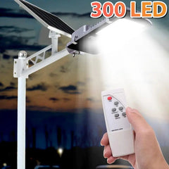A hand holds a remote control in front of a solar-powered LED street light mounted on a pole.