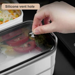 A hand pulls the silicone vent plug on the lid of a rectangular stainless steel lunch box.