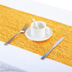 A gold sequin table runner is displayed on a white surface with a cup, saucer, fork, and knife.