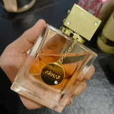 A hand holds a rectangular glass bottle of Armaf Club De Nuit perfume with a gold charm.