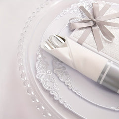 A white plastic plate with silver filigree trim holds a pre-rolled napkin containing silverware.
