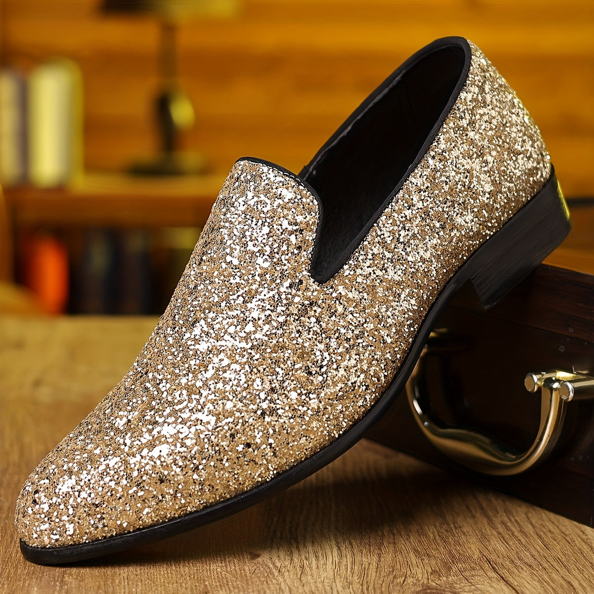 A gold glitter slip-on dress shoe with a pointed toe and black trim sits on a wooden surface.