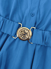 A close-up of a blue dress featuring a gold circular belt buckle with an intricate cutout design.
