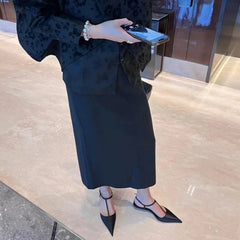 A person wears black pointed-toe t-strap flats with a low heel and a long black skirt.