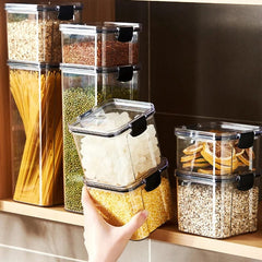A hand holds a clear plastic storage container filled with yellow grains next to a set of jars.