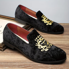 A pair of black velvet slip-on loafers featuring gold embroidered crests on the vamps.