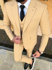 A man wears a tan three-piece suit with a white shirt, black tie, and black slip-on shoes.