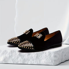 Black velvet loafers with gold spiked toe caps and metallic hardware on a white stone base.