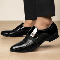 Men's black slip-on dress shoes featuring a pointed toe, embossed pattern, and metallic detail.