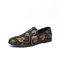 A black slip-on men's shoe featuring a gold floral embroidery pattern and a black rubber sole.