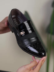 A black slip-on dress shoe with a pointed toe, embossed detailing, and a metallic accent.