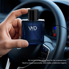 A hand holds a square blue glass bottle of Charm Ocean Eau de Cologne in front of a car steering wheel.