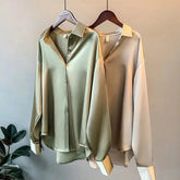 Two long-sleeved silk-style button-down shirts in sage green and champagne hanging on hangers.