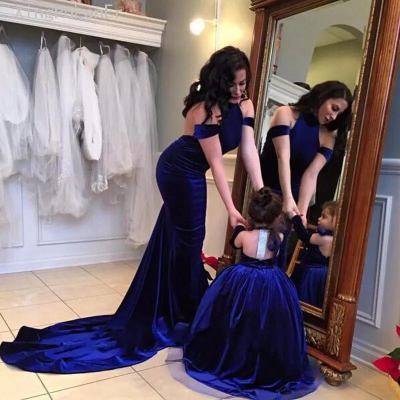 A woman and girl wear matching Backless Velvet Flower Girls Dresses—royal blue, off-the-shoulder ball gowns—admiring their reflection in a dress shop surrounded by elegant first communion dresses.