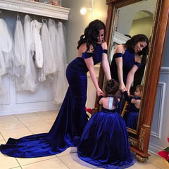 A woman and girl wear matching Backless Velvet Flower Girls Dresses—royal blue, off-the-shoulder ball gowns—admiring their reflection in a dress shop surrounded by elegant first communion dresses.