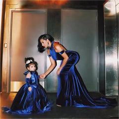 A woman and girl wear matching Backless Velvet Flower Girls Dresses—royal blue, off the shoulder, long ball gowns—standing indoors by metallic gray doors. Elegant hairstyles make these perfect for flower girls or first communion.