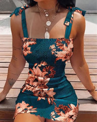 A woman wears a teal, floral-print, sleeveless mini dress with a square neckline and tie straps.
