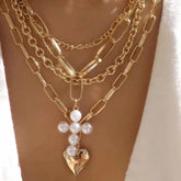 A multilayer gold chain necklace featuring a pearl cross pendant with a heart charm at the base.
