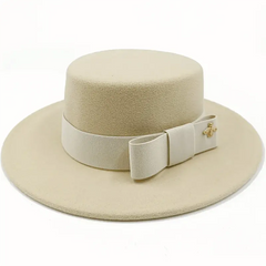 A beige flat-top fedora hat featuring a matching ribbon bow and a small gold bee emblem.