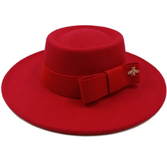A red flat-top fedora hat featuring a matching ribbon bow and a gold bee emblem.