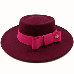 A red felt fedora hat with a flat top, featuring a bright pink ribbon bow and a gold bee pin.