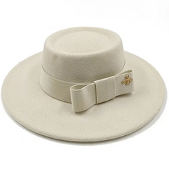 A cream-colored flat-top fedora hat featuring a matching bow and a small gold bee emblem.
