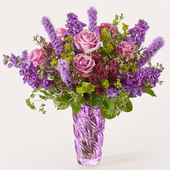 A bouquet of purple roses, stock, and liatris in a textured purple glass vase.