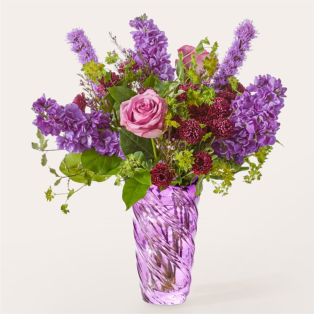 A bouquet of purple roses, stock, and dark red flowers in a textured purple glass vase.