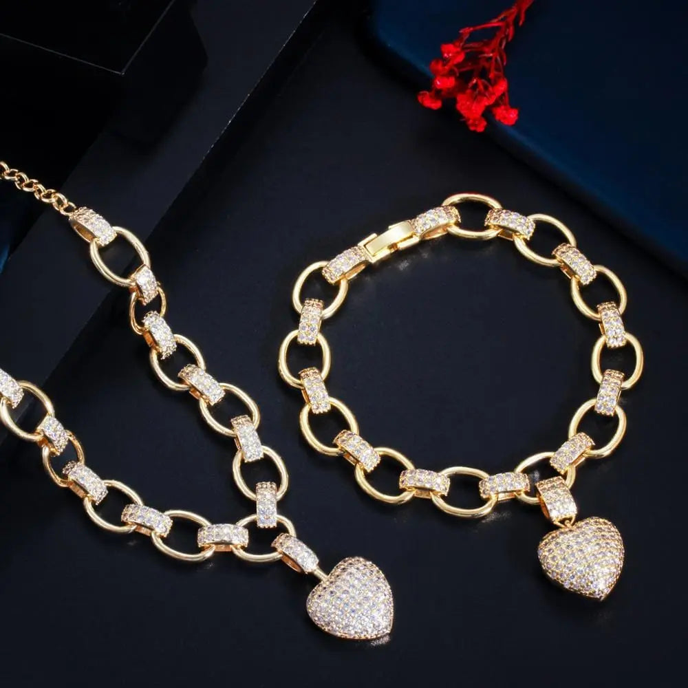 The CWWZircons 585 Gold Color Cubic Zirconia Dangle Love Heart Shape Charm Bracelet and Pendant Necklace Set (T468) features sparkling heart charms, elegantly showcased with a red dried flower on a black surface.