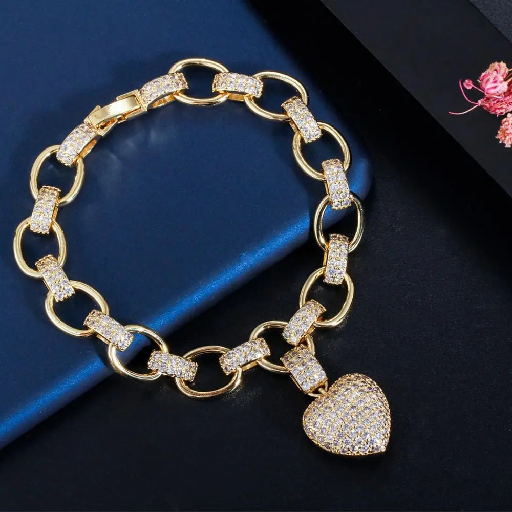 The CWWZircons 585 Gold Color Cubic Zirconia Love Heart Charm Bracelet from set T468 features oval links and a sparkling heart pendant, displayed on a dark textured surface with a blue item and floral decoration.