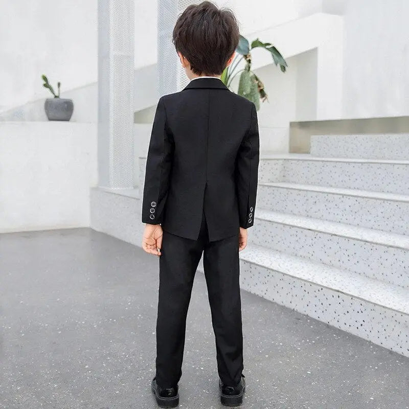 A young boy wears the Children Formal Dress Kids Flower Jacket Pants Bowtie 3PCS Photography Suit, standing indoors with his back to the camera, facing a white staircase as a potted plant sits on a nearby ledge.