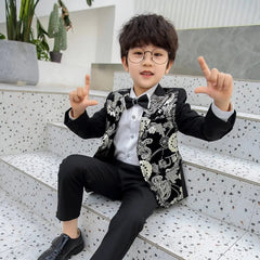 A young boy in a Children Formal Dress Kids Flower Jacket Pants Bowtie 3PCS Piano Costume—a stylish black bronzing show host photography suit—smiles on terrazzo stairs, making an L shape with both hands.