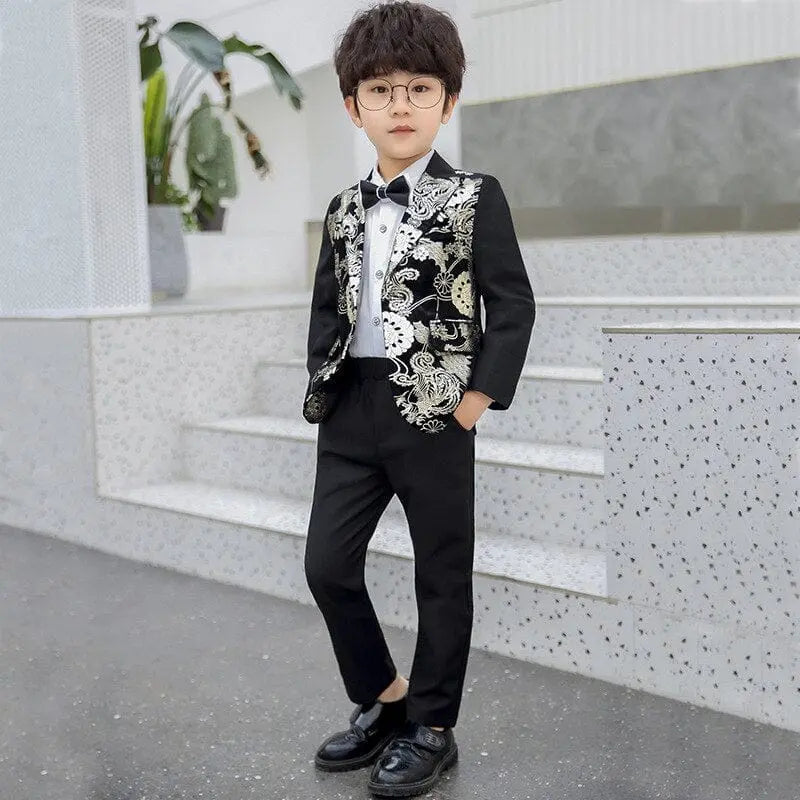 A young boy with glasses stands confidently on indoor steps, wearing the Children Formal Dress Kids Flower Jacket Pants Bowtie 3PCS—a handsome photography suit featuring a black outfit and a gold bronzing flower jacket, with his hands in his pockets.