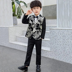A young boy wearing glasses and a black suit with an ornate silver floral pattern stands confidently outdoors, modeling the Children Formal Dress Kids Flower Jacket Pants Bowtie 3PCS Piano Costume Boys Bronzing Show Host Handsome Photography Suit.